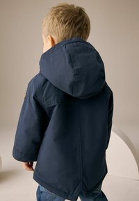 Veste imperméable bleu marine avec capuche, présentant une texture lisse et des détails de couture subtils, portée avec un jean en denim.