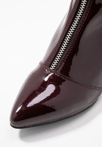 Bottine en cuir verni bordeaux avec un bout pointu, dotée d'une fermeture éclair argentée sur le côté et d'une texture lisse et brillante.