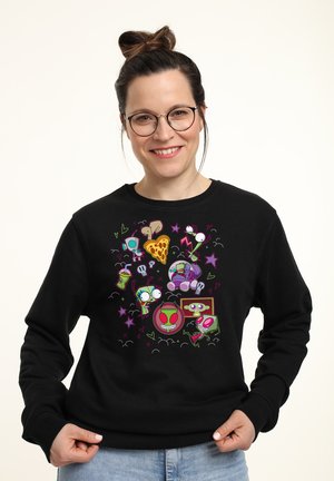 Femme avec des lunettes et un chignon, portant un sweat-shirt noir avec des extraterrestres colorés de dessin animé, une part de pizza, des étoiles et des illustrations de boissons.