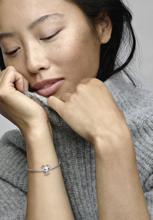 Bracelet en argent avec un charm représentant un éléphant et des accents roses, porté sur une main devant un fond de pull gris texturé.