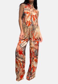 Jumpsuit rosso con stampa tropicale e collo a halter, con dettaglio di frange in vita. Il tessuto ha una consistenza liscia e pantaloni larghi.