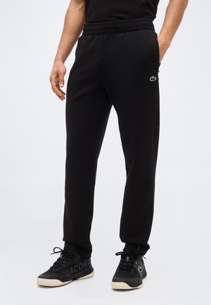 Άνδρας που φοράει μαύρο παντελόνι φόρμας με το λογότυπο της Lacoste στην τσέπη και μαύρα αθλητικά παπούτσια Lacoste με μπεζ σόλες, στέκεται μπροστά σε απλό φόντο.