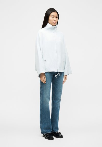 Γυναίκα που στέκεται με ανοιχτό μπλε μπουφάν με ψηλό γιακά, μπλε τζιν με ίσια μπατζάκια και μαύρα δερμάτινα παπούτσια σε λευκό φόντο.