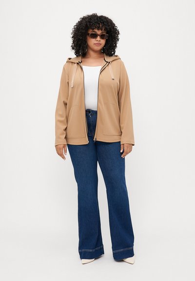 Haut de survêtement beige zippé avec capuche à cordon, deux poches avant, porté sur un haut blanc et un jean évasé bleu foncé, associé à des lunettes de soleil.
