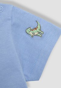 Svijetloplava pamučna majica s kratkim rukavima. Ima izvezenog zelenog dinosaura s crvenim točkicama na lijevom rukavu.