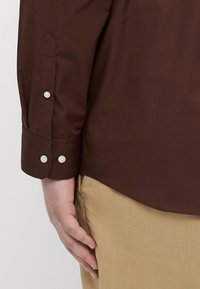 Brun langermet skjorte med knappede mansjetter, som har hvite knapper og et glatt, teksturert stoff. Kombinert med lyse beige bukser.