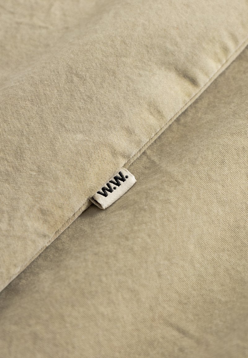 Coussin en tissu beige à surface texturée, avec une petite étiquette cousue portant la mention « W.A.N. » près du bord.