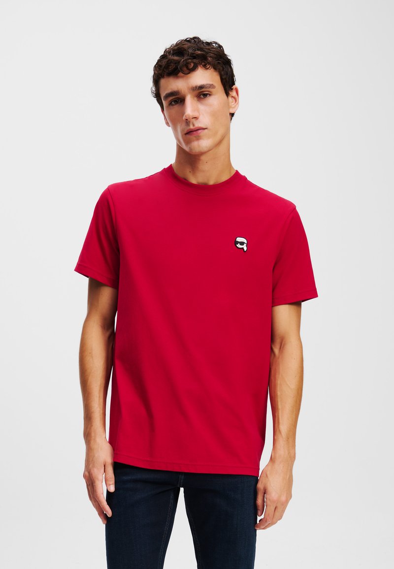 Tricou roșu din bumbac, cu decolteu rotund și mâneci scurte, având un mic logo stil emoji de culoare neagră în zona stângă a pieptului.