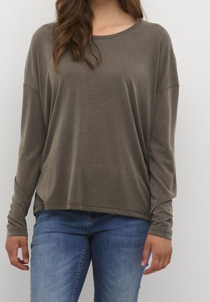 Personne portant une chemise ample à manches longues gris-brun et un jean bleu, debout devant un fond clair uni.