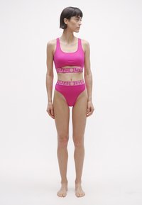 Ružové dvojdielne plavky pozostávajúce z krátkeho topu s výrezom a nohavičiek s vysokým pásom, oboje zdobené zlatým grécko-kľúčovým vzorom.