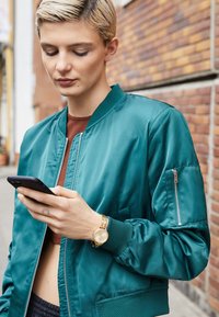 Teal bomber jacket with a satin finish, zippered pockets, and ribbed cuffs, paired with a gold watch, while holding a smartphone.