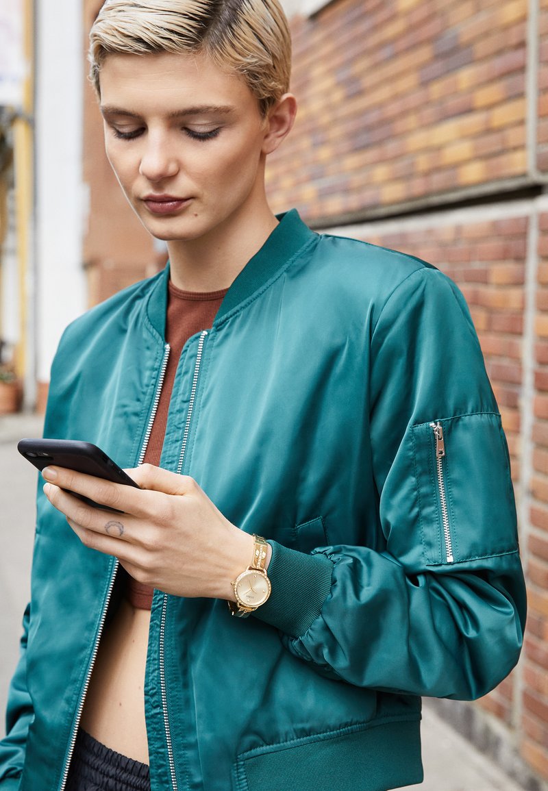 Teal bomber jacket with a satin finish, zippered pockets, and ribbed cuffs, paired with a gold watch, while holding a smartphone.