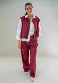 Vrouw staand tegen een effen achtergrond, gekleed in een wit shirt, bordeauxrode textuurvest en bijpassende broek, met witte sneakers en gouden oorringen.