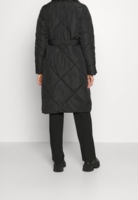 Zwarte gewatteerde jas met een diamantpatroon, een ceintuurlengte en lange mouwen. Draagt over zwarte broeken en flinke zwarte laarzen.