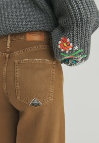 Brune denimbukser med et trekantet logo-patch, parret med en struktureret grå sweater med farverig blomsterbroderi på ærmet.