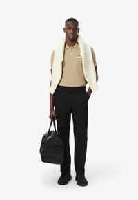 Beige polo-shirt med grön accent, vit stickad tröja över draperad, svarta byxor, svart läderväska, kraftiga svarta sneakers, neutral bakgrund.
