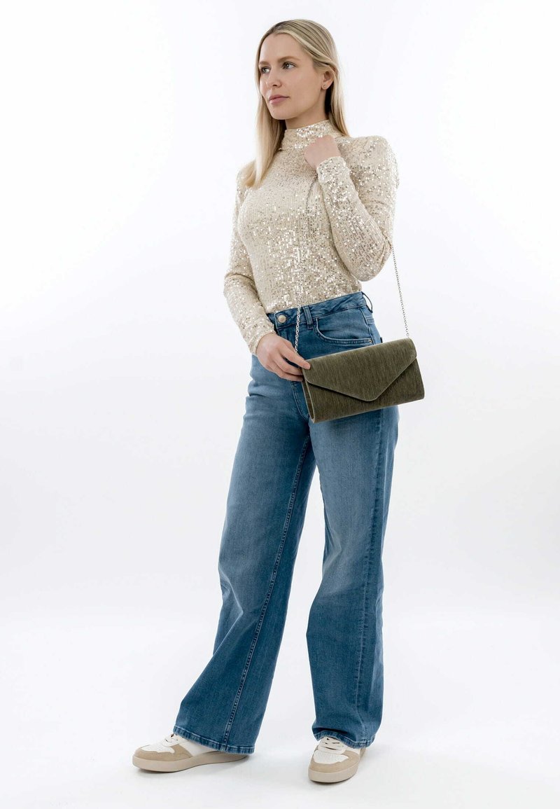 Top a maniche lunghe in beige con paillettes, abbinato a jeans blu a vita alta e gamba larga. Tiene in mano una borsa a busta in velluto verde con tracolla a catena.
