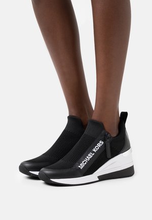 Black slip-on sneakers with mesh texture and a zip detail, featuring a white logo and a wedge sole. Accented with glossy black and white elements.