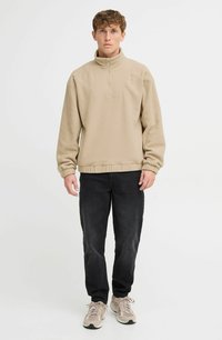 Beige fleece pullover med halv lynlås krave, der har et tekstureret panel, ribbede ærmer og kant, sammen med mørke lige ben jeans.