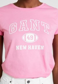 Osoba v světlém růžovém tričku s bílým nápisem "GANT", ikonou fotbalu s číslem "49" a nápisem "NEW HAVEN", v kombinaci s bílými kalhotami.