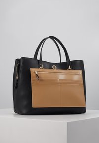 Sac fourre-tout en cuir noir avec une poche avant beige, doté de quincaillerie dorée et de deux poignées. Surface texturée avec un détail de fermeture éclair.
