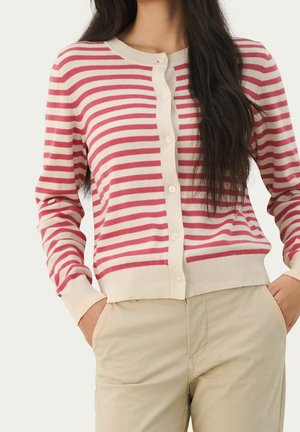 Femme portant un cardigan boutonné à rayures horizontales roses et blanches avec un pantalon beige, mains dans les poches, longs cheveux foncés visibles.