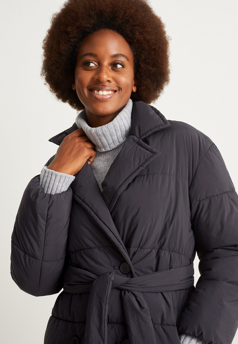 Eine lächelnde Frau mit lockigem Haar, die einen dunklen, taillierten Daunenmantel über einem hellgrauen Rollkragenpullover trägt und zur Seite schaut.