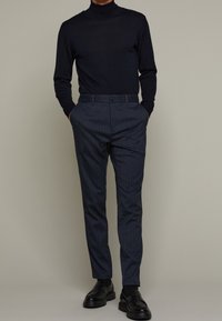 Pull à col roulé foncé associé à un pantalon gris à fines rayures. Le pantalon présente une coupe classique et des poches avant. Chaussures : mocassins noirs.