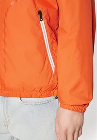 Primo piano di una persona che indossa una giacca arancione con tasca laterale con zip e polsini elasticizzati, abbinata a jeans azzurri chiari.