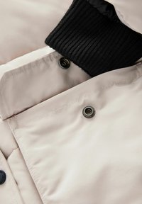 Hellbeige Jacke mit einem schwarzen Rippkragen und Knopfverschluss, die eine glatte Textur und minimalistisches Design aufweist.