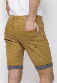 Rjavi jeans kratke hlače s zavitki, ki imajo zadnje žepe in svetel etiket na pasu, ter prikazujejo gladko, teksturirano blago.