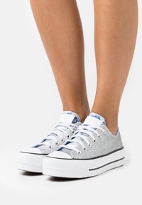 Stříbrné třpytivé tenisky s nízkým designem, bílé gumové podrážky a modrými vzorovanými detaily na jazyku. Šněrovány bílými tkaničkami.