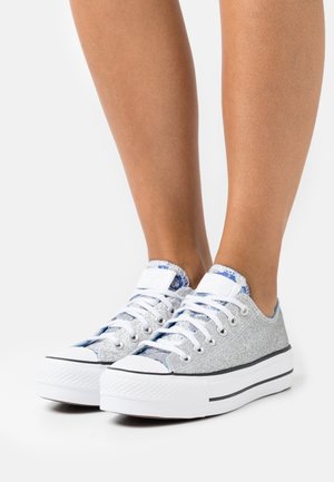 Srebrne lśniące trampki z niskim krojem, białą gumową podeszwą i niebieskimi wzorami na języku. Sznurowane białymi sznurówkami.