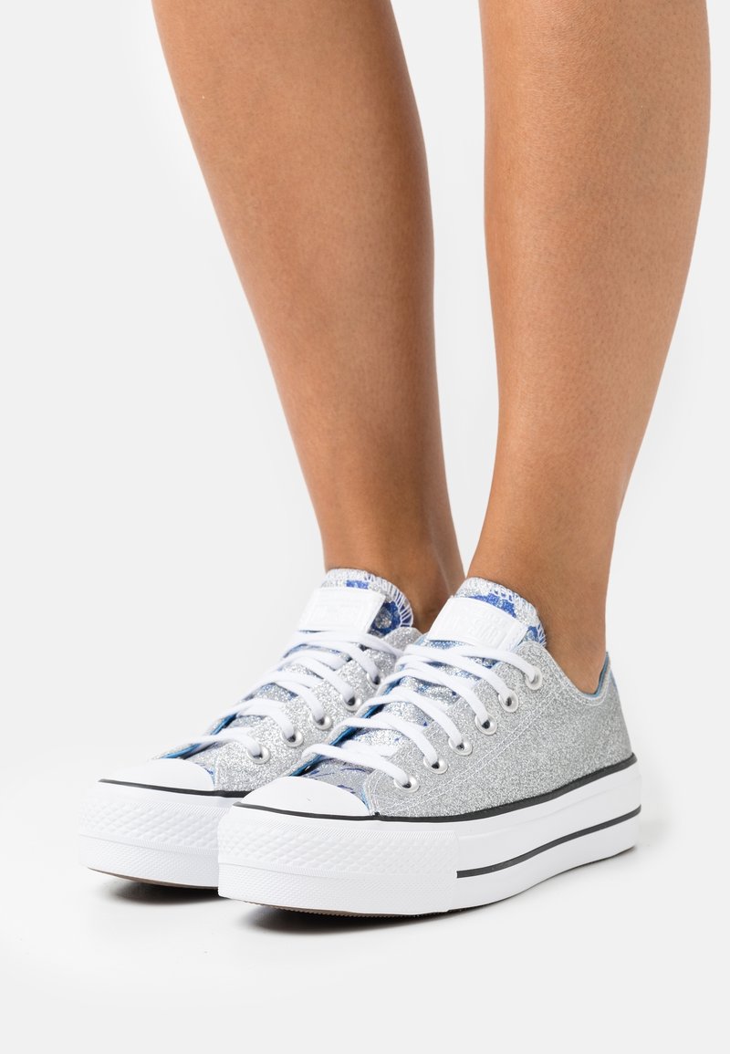 Stříbrné třpytivé tenisky s nízkým designem, bílé gumové podrážky a modrými vzorovanými detaily na jazyku. Šněrovány bílými tkaničkami.