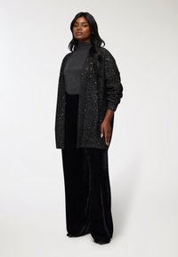 Kobieta stoi w ciemnoszarej golfie, czarnym świecącym kardiganie i szerokich czarnych welurowych spodniach na tle jasnego, jednolitego tła.