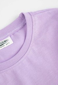 Fioletowa bawełniana koszulka z okrągłym dekoltem, prążkowanym wykończeniem i starannym przeszyciem. Widoczna metka z informacjami o rozmiarze i marce.