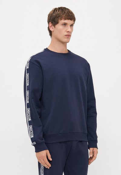 Ung mann iført en marineblå hettegenser og matchende bukser med hvite Moschino-logo striper på ermene og buksebeina, står mot en hvit bakgrunn.