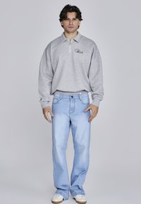 Felpa grigia con zip e colletto e logo, abbinata a jeans azzurri a gamba dritta e sneakers bianche su uno sfondo neutro.