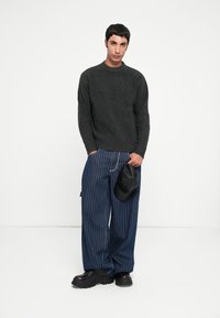 Dark gray knitted sweater with a round neckline, paired with blue pinstriped wide-leg pants. Model holds a black cap in one hand. Black chunky shoes.