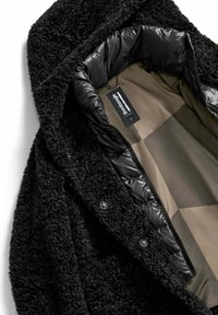 Zwarte fleece buitenlaag met een pluizige textuur en een glanzende zwarte puffer voering. Beschikt over een kap, ritsluiting en drukknoopsluiting.
