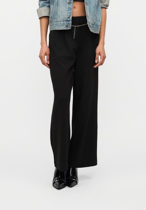 Osoba nosiaca skrátenú džínsovú bundu a čierne široké nohavice s vysokým pásom spolu s čiernymi čižmami s špicatou špičkou, stojaca na bielom pozadí.