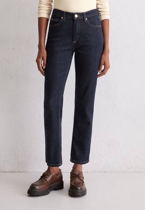 Dunkelblaue Jeans aus Denim mit geradem Beinausschnitt, ausgestattet mit Gürtelschlaufen, Vorderentaschen und kontrastierendem orangem Nähen. Kombiniert mit braunen Schuhen.