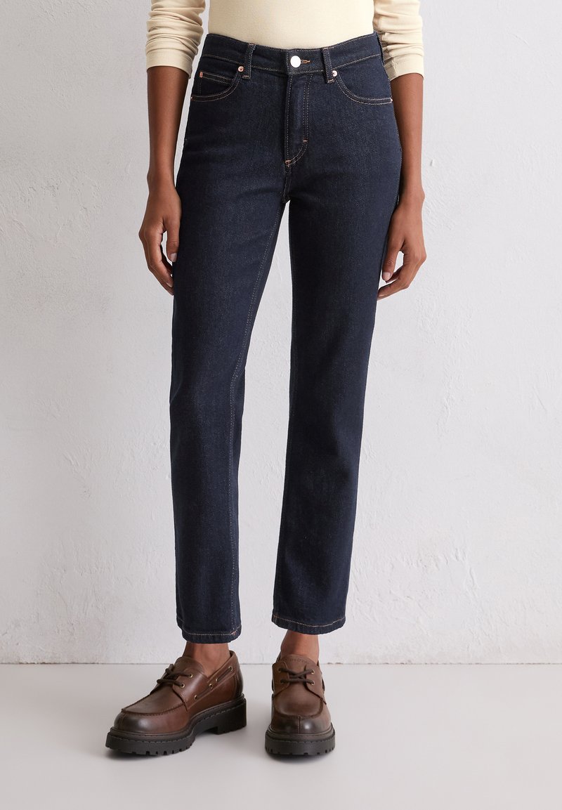 Donkerblauwe spijkerbroek met een rechte pijp, voorzien van riemlussen, zijzakken en contrasterende oranje stiksels. Geshstyled met bruine schoenen.