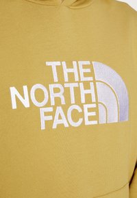 Sweatshirt amarelo com capuz, apresentando um grande logotipo branco bordado "THE NORTH FACE" com um gráfico de destaque curvado. Tecido suave e texturizado.