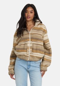 Giacca a zip fuzzy in tonalità talpa, beige e crema con motivo a strisce. Presenta un colletto alto, tasche laterali e polsini a costine. Tessuto morbido e testurizzato.