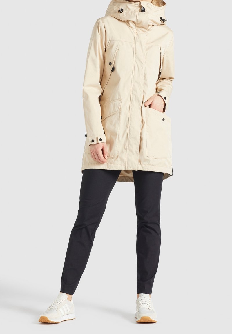 Beżowa wodoodporna parka z kapturem, dwoma przednimi kieszeniami i czarnymi guzikami. Nałożona na ciemne spodnie i białe trampki. Gładka tekstura.