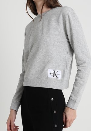 Sudadera gris recortada hecha de tejido suave, con cuello redondo y un parche con el logo en la parte frontal. Diseño simple y minimalista.