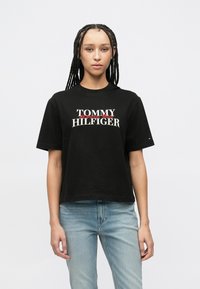 Μαύρο βαμβακερό T-shirt με το λογότυπο "TOMMY HILFIGER" σε λευκό με κόκκινες λεπτομέρειες. Κοντά μανίκια και άνετη ε�φαρμογή, με μικρό λογότυπο στο μανίκι.