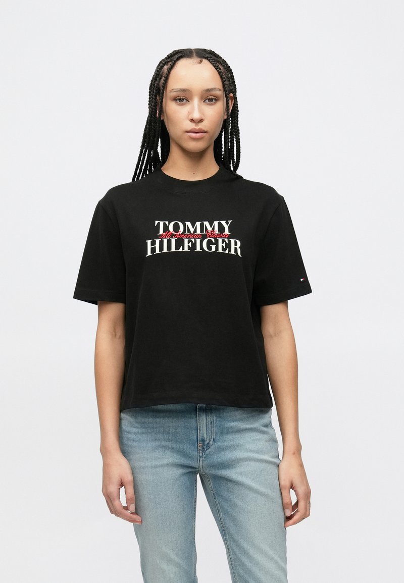 Μαύρο βαμβακερό T-shirt με το λογότυπο "TOMMY HILFIGER" σε λευκό με κόκκινες λεπτομέρειες. Κοντά μανίκια και άνετη εφαρμογή, με μικρό λογότυπο στο μανίκι.