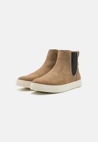 Bottes courtes en daim marron avec panneaux élastiques sur les côtés et semelles blanches en caoutchouc. Texture lisse avec un bout arrondi et un design minimaliste.